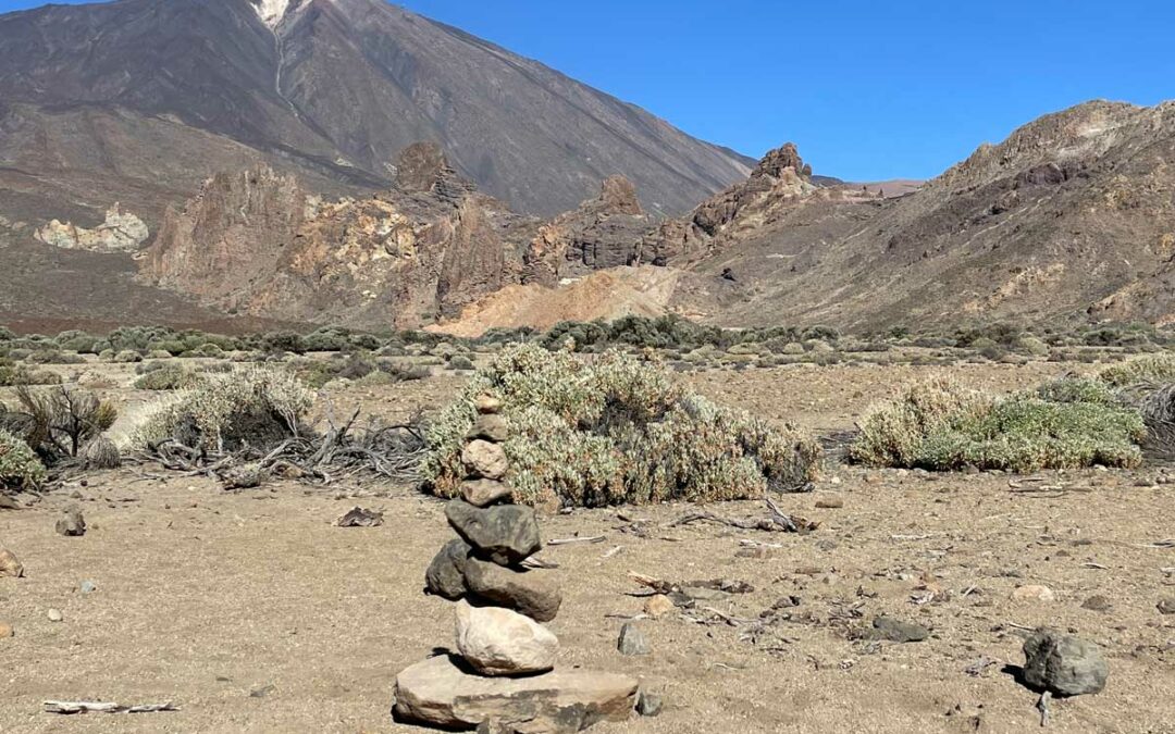 Ein von Menschen aufgeschichtetes Steintürmchen steht in der Ucana Ebene des Teide Nationalparks. Der Steintrum ist umgeben vom typischen Teide-Ginster und im Hintergrund ist die Felsformation Roques de Garcia und der Teide zu sehen.