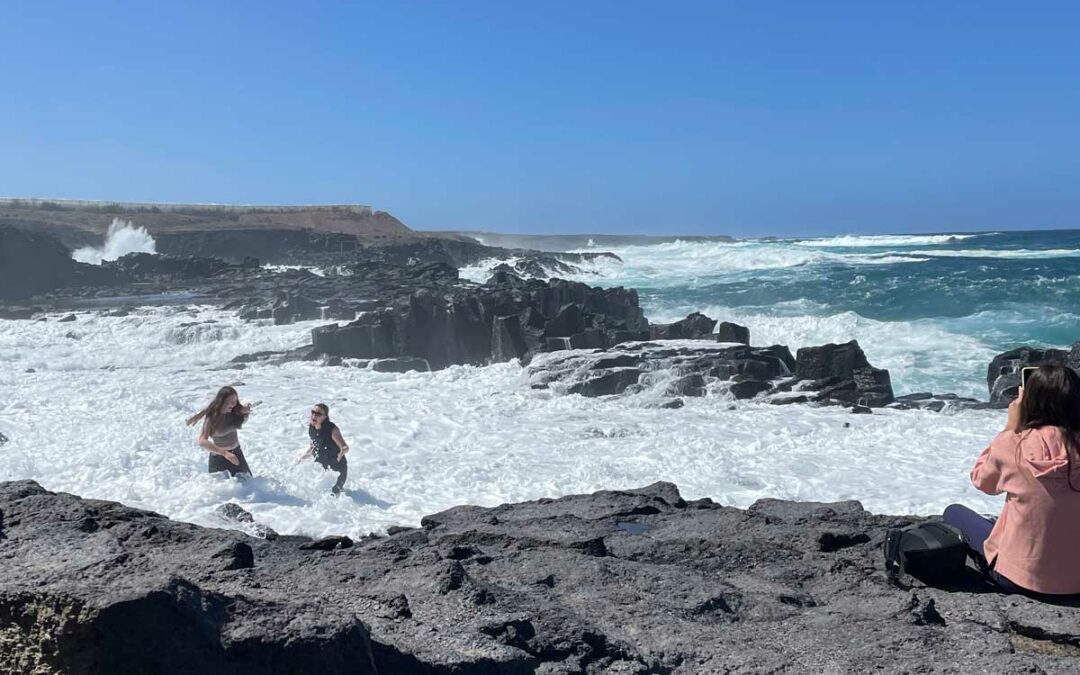 An der Küste fotografiert eine Frau das stürmische Meer, während zwei weitere Frauen von hohen Wellen überrascht und beinahe ins Meer gespült werden.